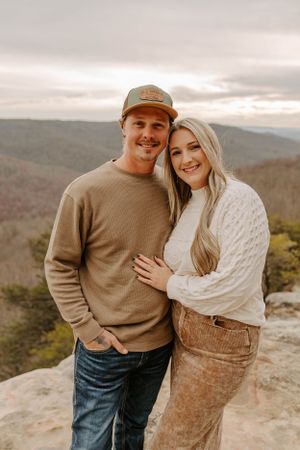 A couple in matching earth-toned sweaters embraces on a rocky overlook with mountains in the background.