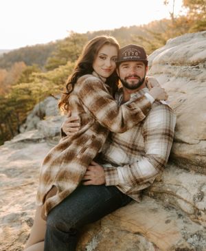 A cozy embrace shared on rocky terrain with fall foliage in the background.