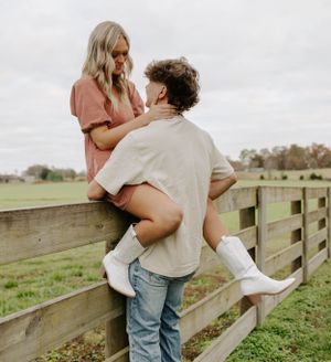 A couple poses romantically on a wooden fence in a countryside setting.