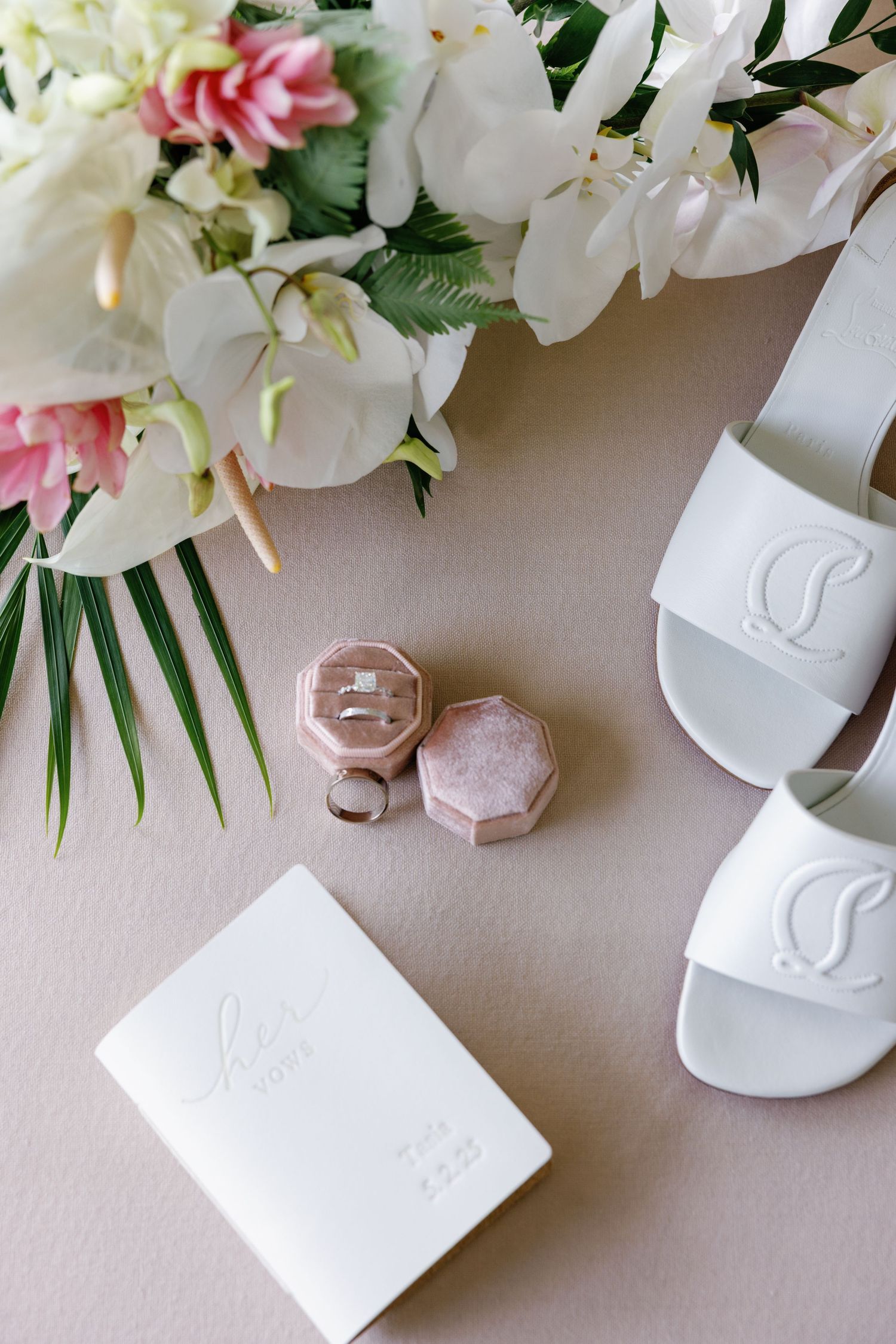 Wedding flat lay with rings in pink velvet box, white heels, vow book, and tropical flowers.