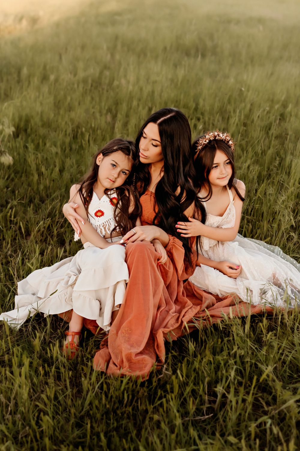 Three people in coordinating dresses sitting in tall grass at sunset.
