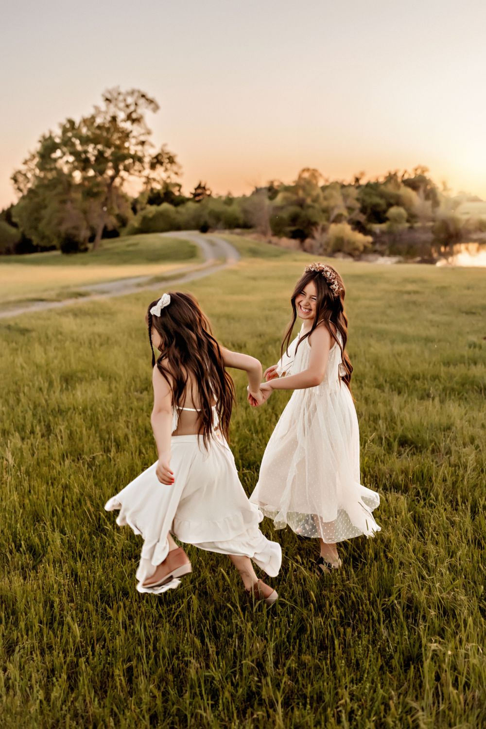 Two women in flowing white dresses dance together in a grassy field at sunset.
