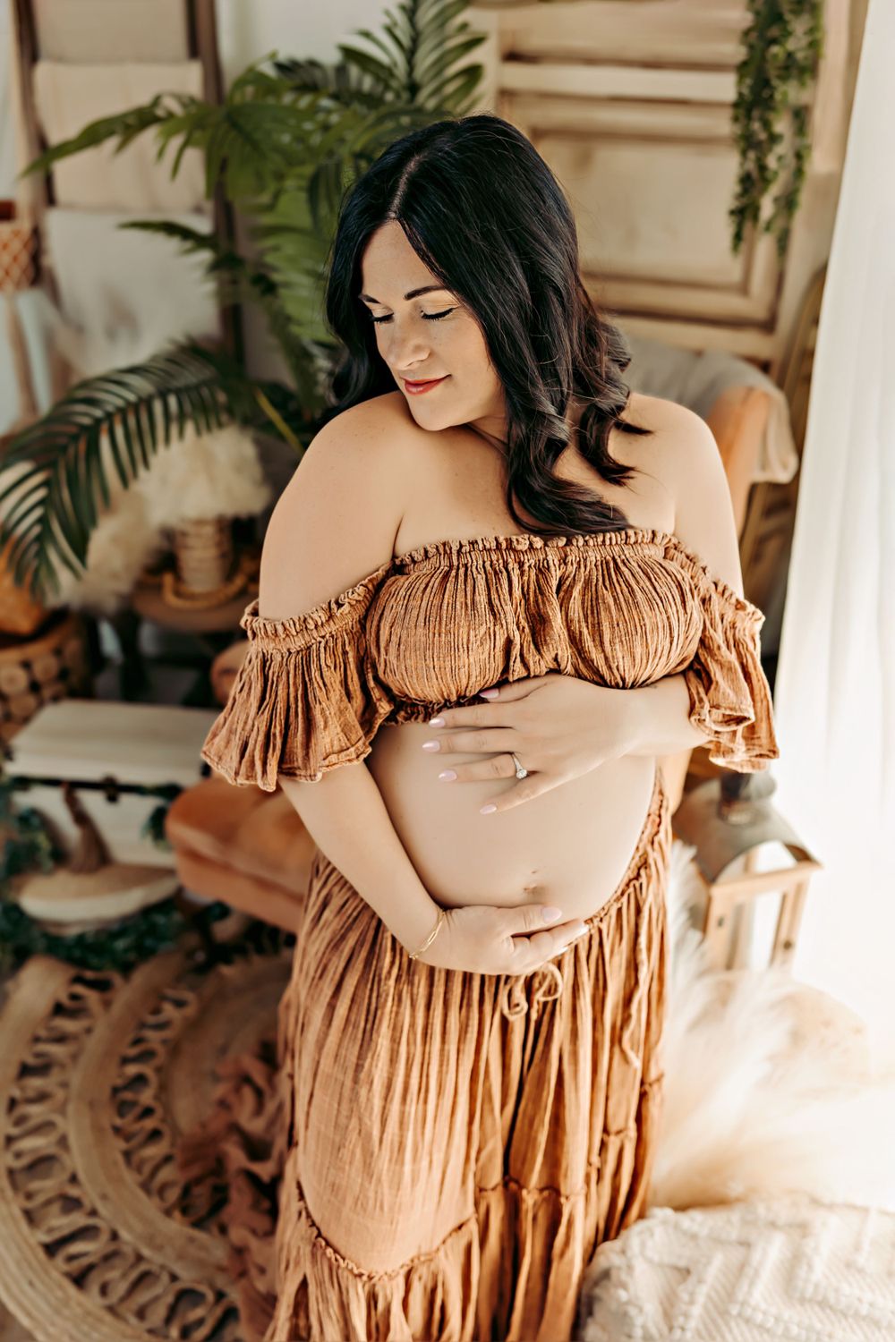 Expectant mother in rust-colored off-shoulder dress poses in bohemian styled room.