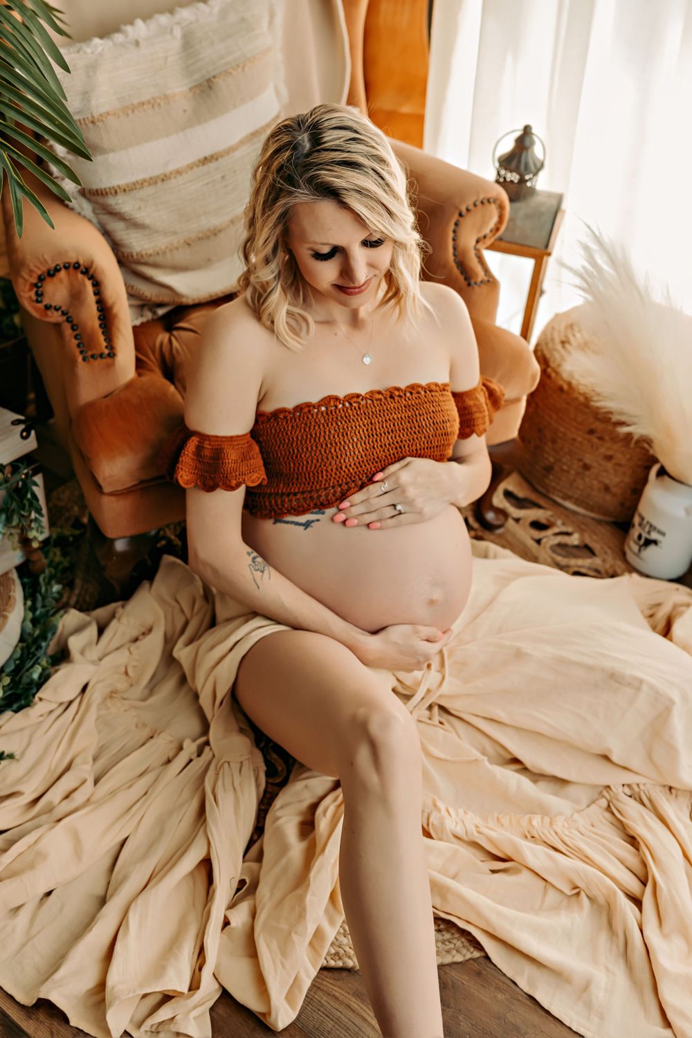 Expectant mother in rust-colored dress poses on beige chaise with boho decor.