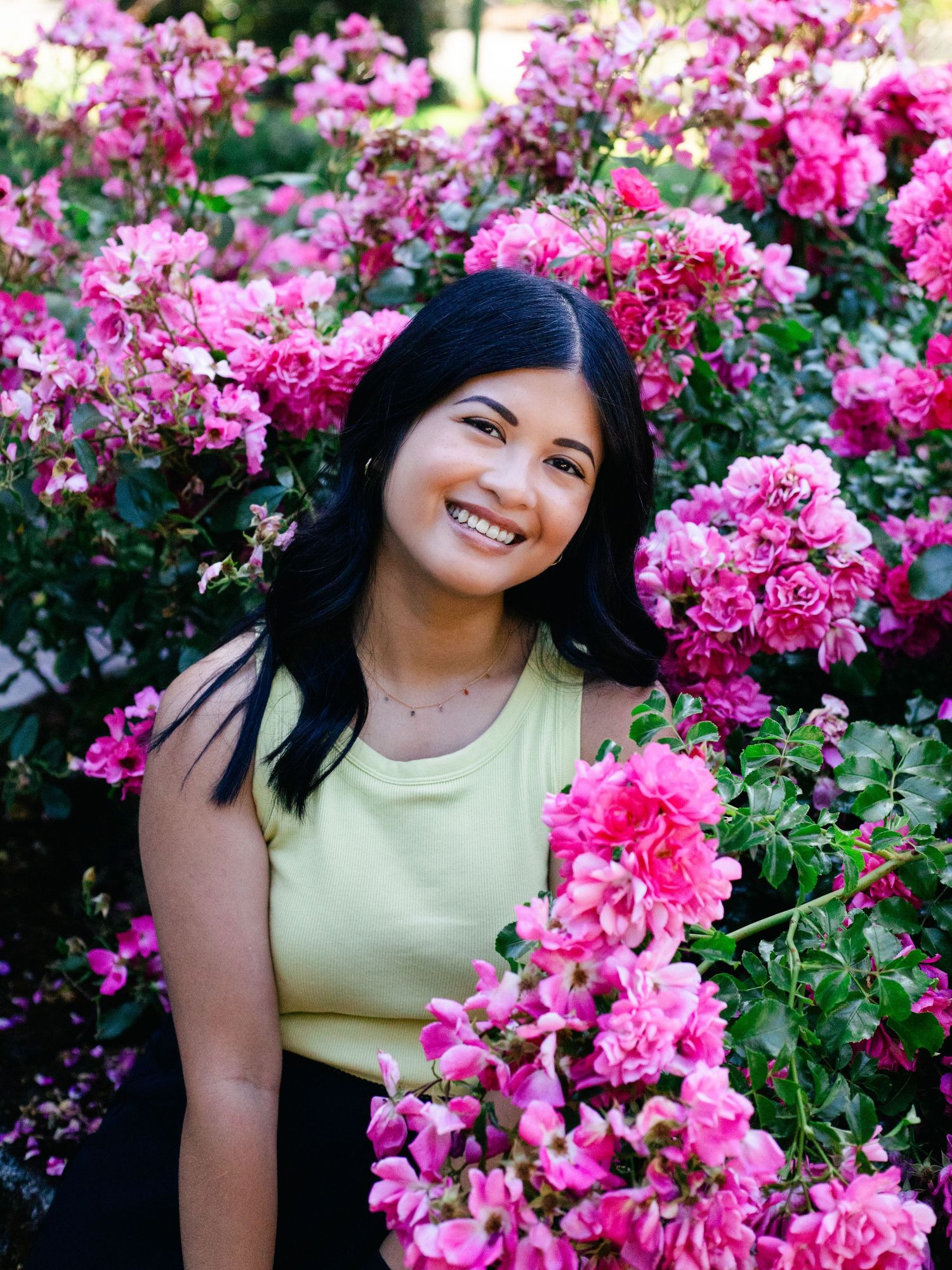 A smiling woman sitting in front of a pink rose bush.