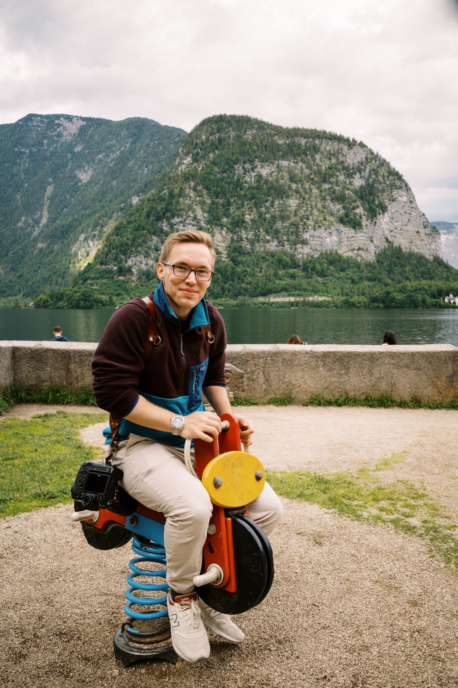 Tourist sits on orange toy motorcycle against backdrop of mountain lake landscape.