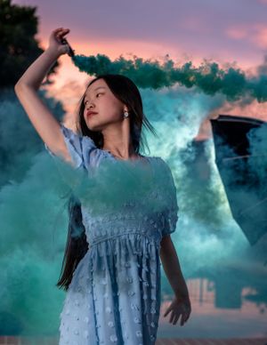 Artistic portrait with turquoise smoke effects and dramatic sunset sky backdrop.
