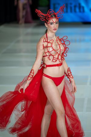 Model wearing avant-garde red swimwear with sculptural wire details and flowing tulle cape on fashion runway.