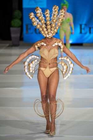 Model on runway wearing avant-garde beige bodysuit with pearl-like embellishments and dramatic crown-shaped headpiece.