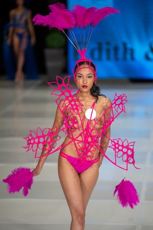 Model walks runway in hot pink swimsuit with dramatic feathered headpiece and geometric cutout accessories at fashion show.
