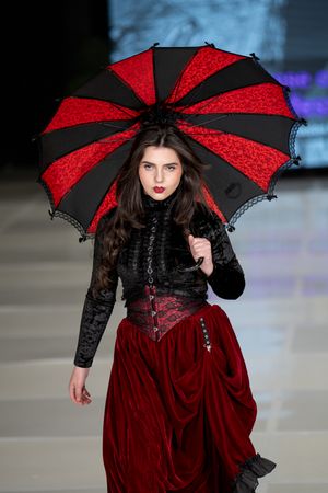 Gothic fashion model on runway wearing black and red Victorian-inspired dress with matching striped parasol umbrella.