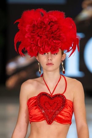 Fashion model walking runway in bright red halter top with heart detail and dramatic red feathered headpiece.