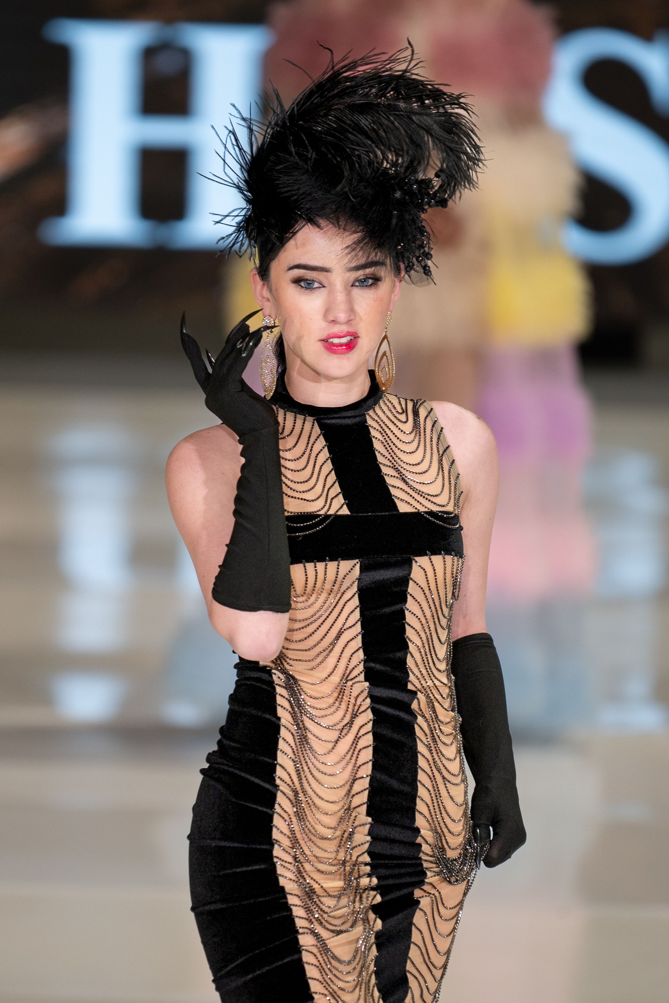 Fashion model in a black and beige gown with a dramatic feather headpiece walks the runway in elegant evening wear.