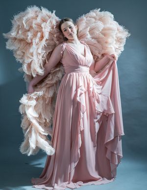 Figure in flowing pink gown stands against dark backdrop with dramatic ruffled feather wings in blush tones.