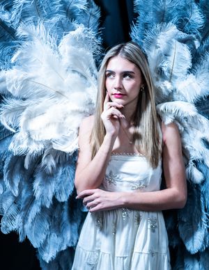 Portrait against a backdrop of blue feathers features a person in a white dress striking a thoughtful pose.