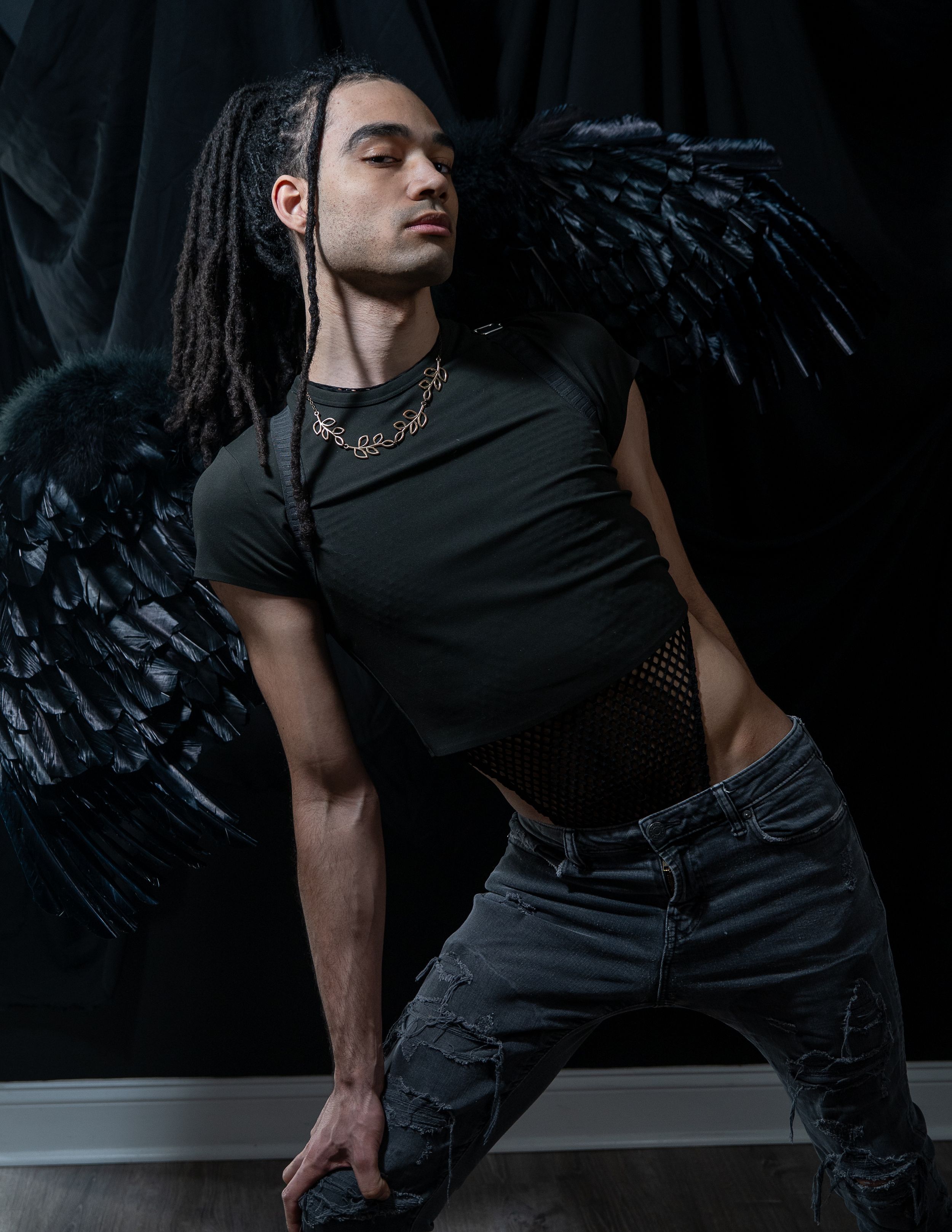 Figure in black outfit with dreadlocks poses dramatically against dark backdrop with black angel wings.