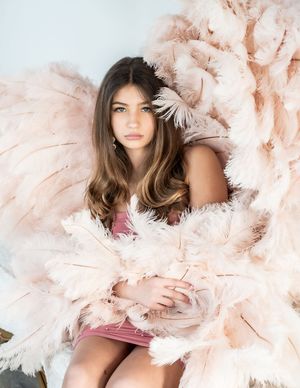 A dreamy fashion portrait surrounded by soft blush pink feathers and plumes creating an ethereal atmosphere.