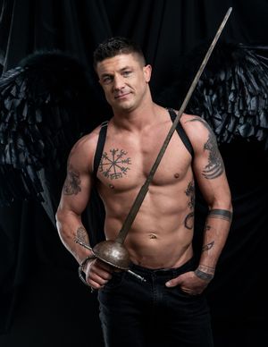 Muscular figure with black angel wings poses shirtless against dark backdrop holding a sword, showing Norse-inspired tattoos.