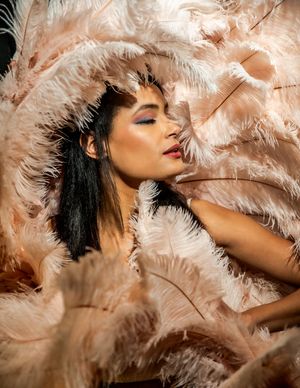 Artistic portrait with soft peach-colored feathers creating a dreamy, ethereal mood against dark hair and glowing skin.