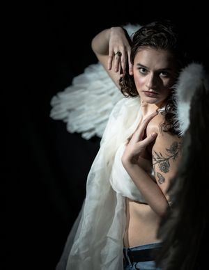 A dramatic dark portrait shows an ethereal winged figure in white fabric with tattoos against a black background.