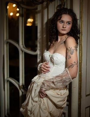 Person wearing a vintage-inspired white corset wedding dress with tattoos poses by ornate doorway in dramatic lighting.