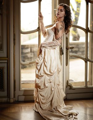 Woman in vintage-style cream dress poses elegantly by large French doors in dramatic lighting.