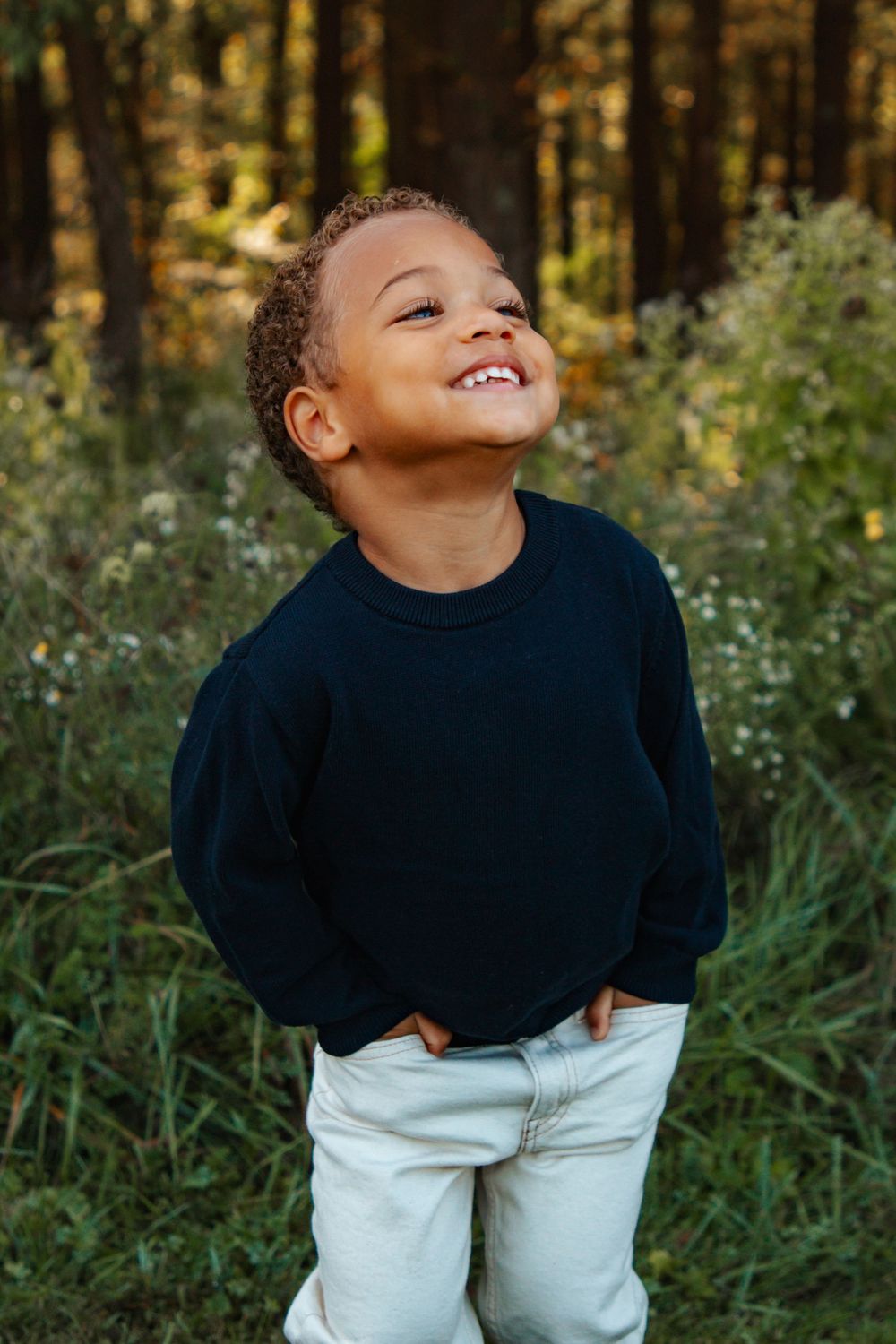 Young child wearing navy sweater and light jeans smiles joyfully while playing outdoors in a wooded setting.