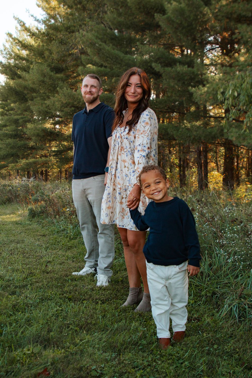 A family stands together in a grassy outdoor setting with evergreen trees during golden hour.