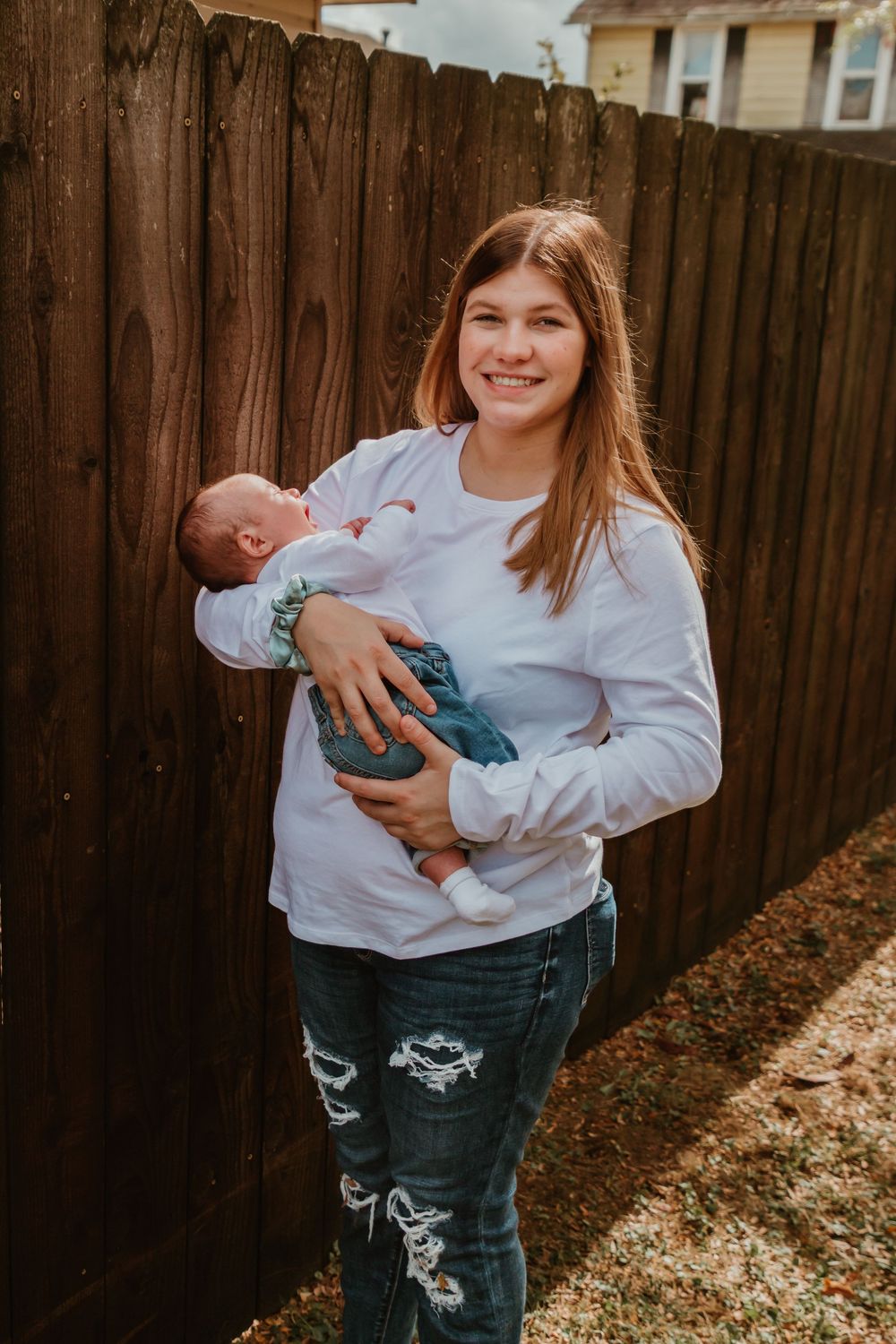 A woman in a white sweater and ripped jeans holds a sleeping newborn baby in front of a wooden fence.