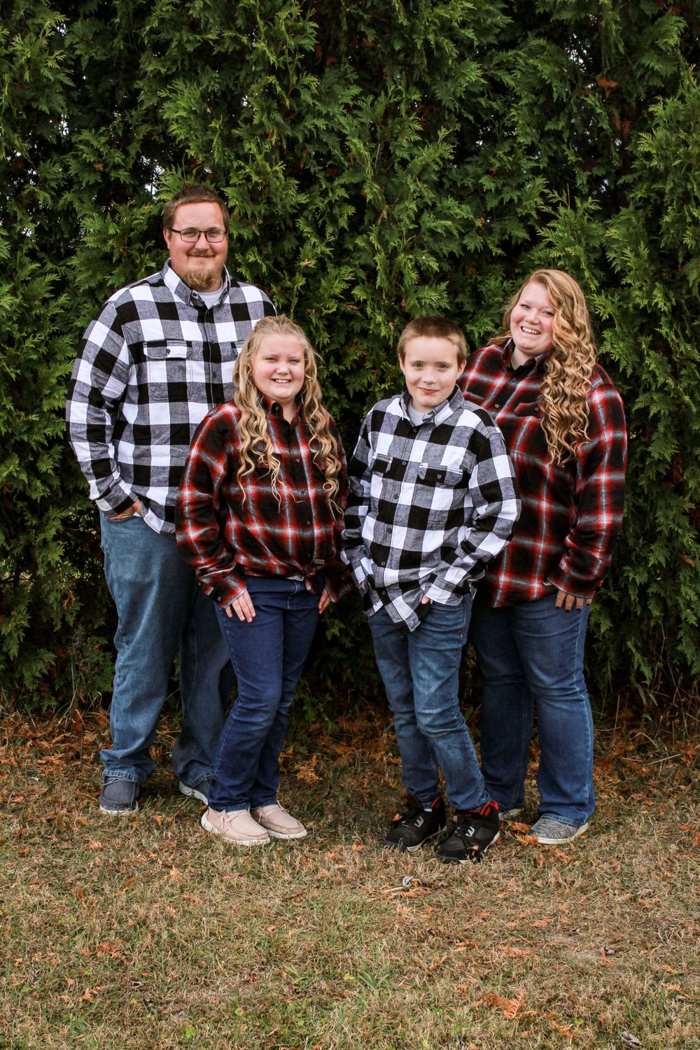 Family of four wearing matching buffalo plaid shirts posing against evergreen trees.