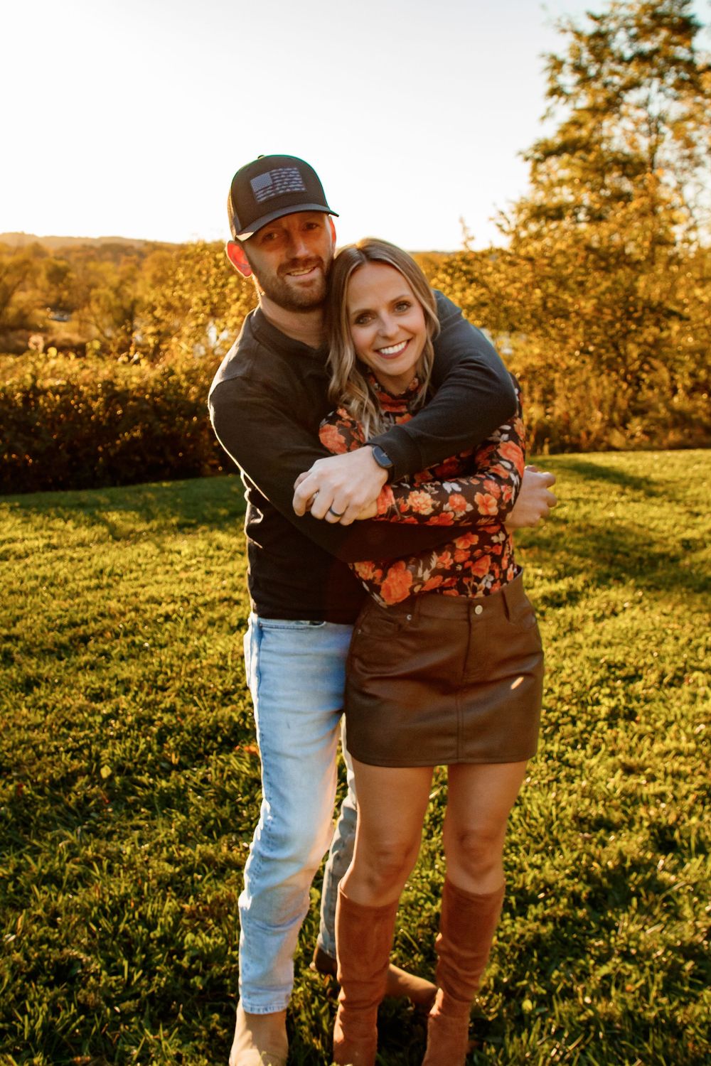 A couple embraces in warm golden sunlight on grassy hillside during autumn sunset.