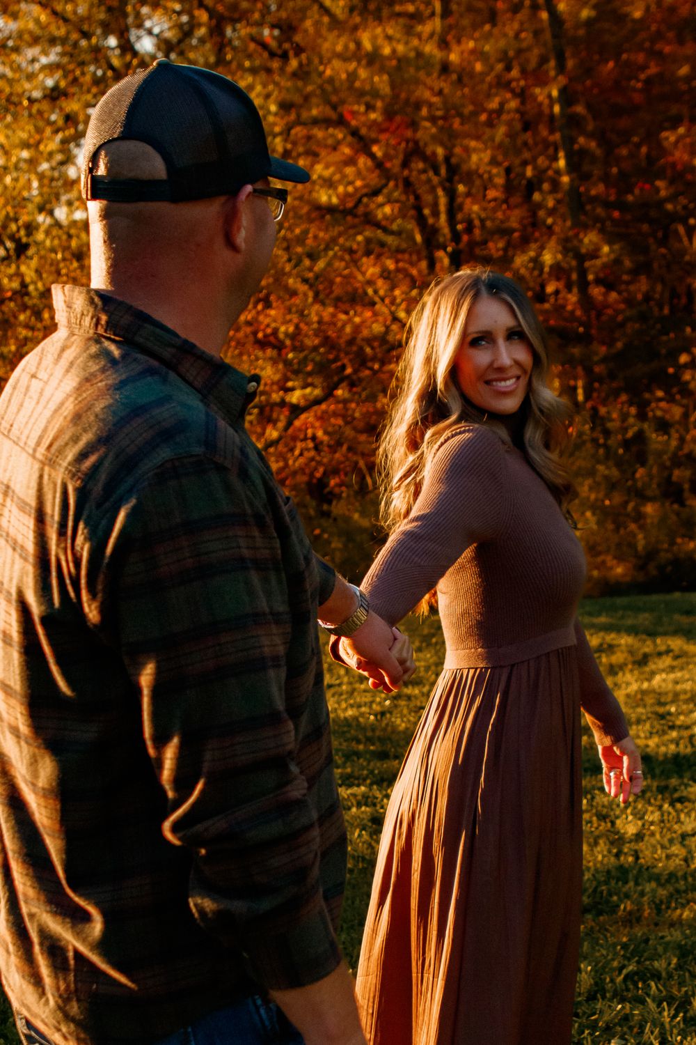 A couple stands together in golden autumn sunlight with fall foliage creating a warm backdrop in the park.