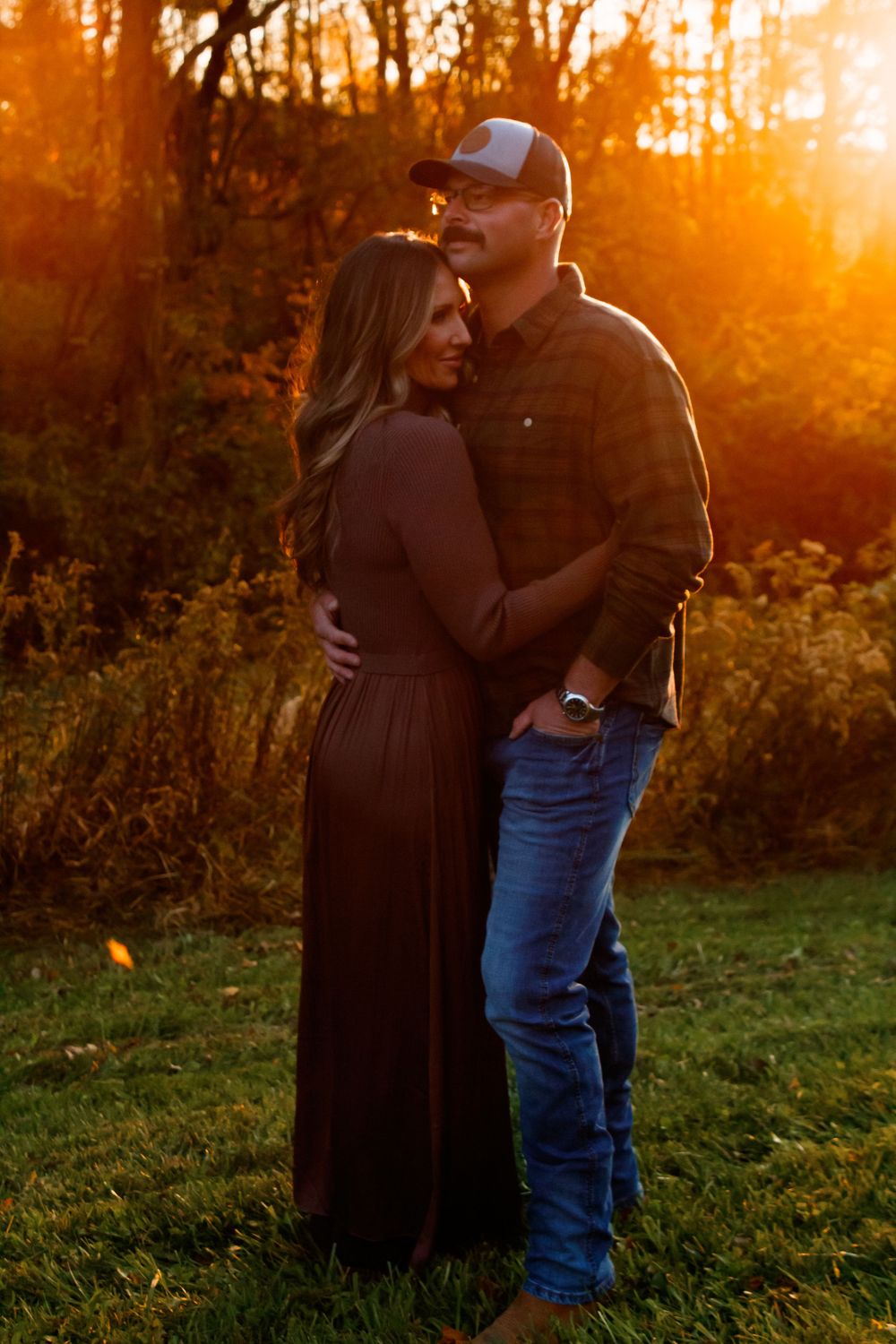 A couple embraces in a romantic outdoor setting during golden hour sunset, silhouetted against warm orange light.