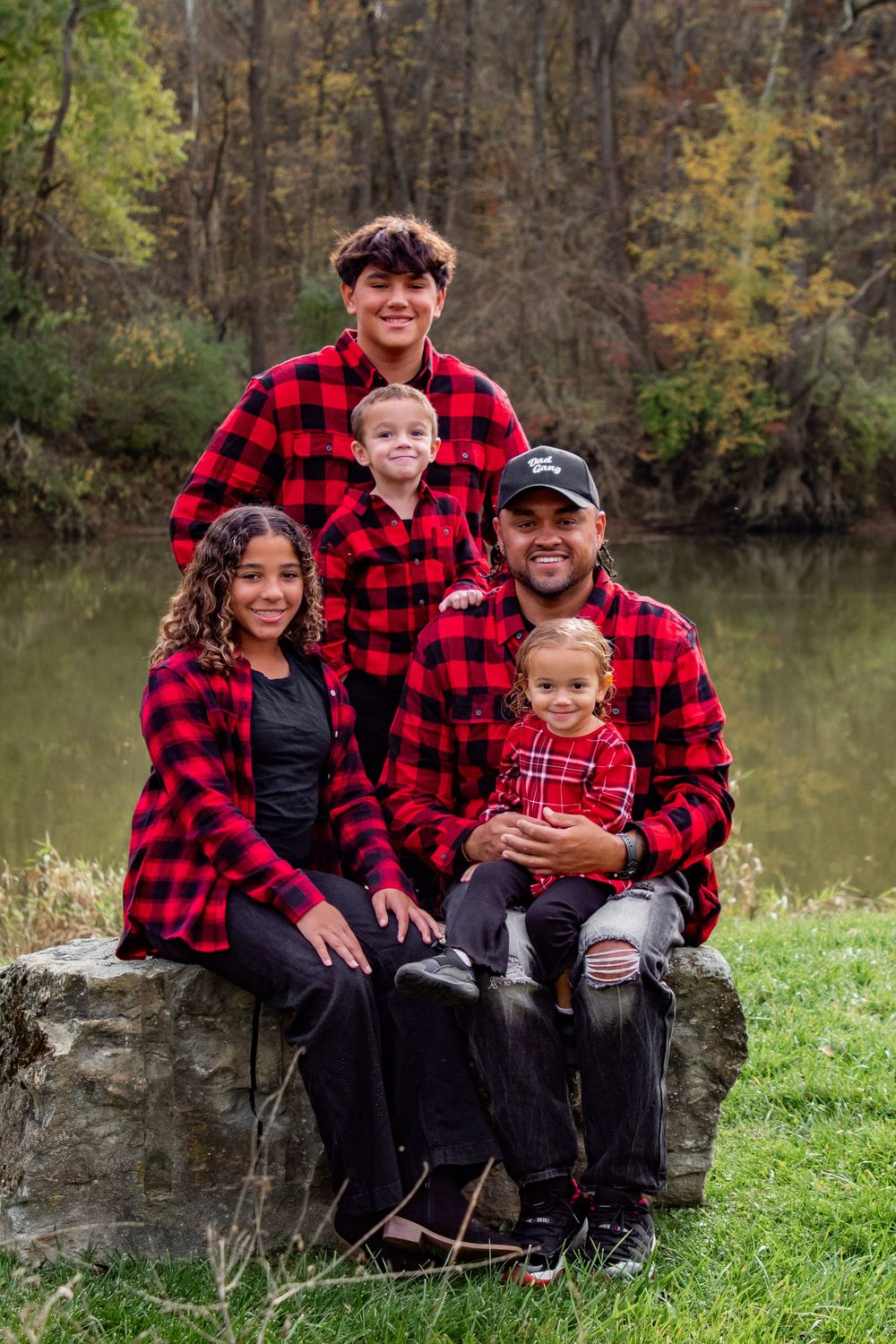A family wearing matching red buffalo plaid flannel shirts poses together by a lake on an autumn day.