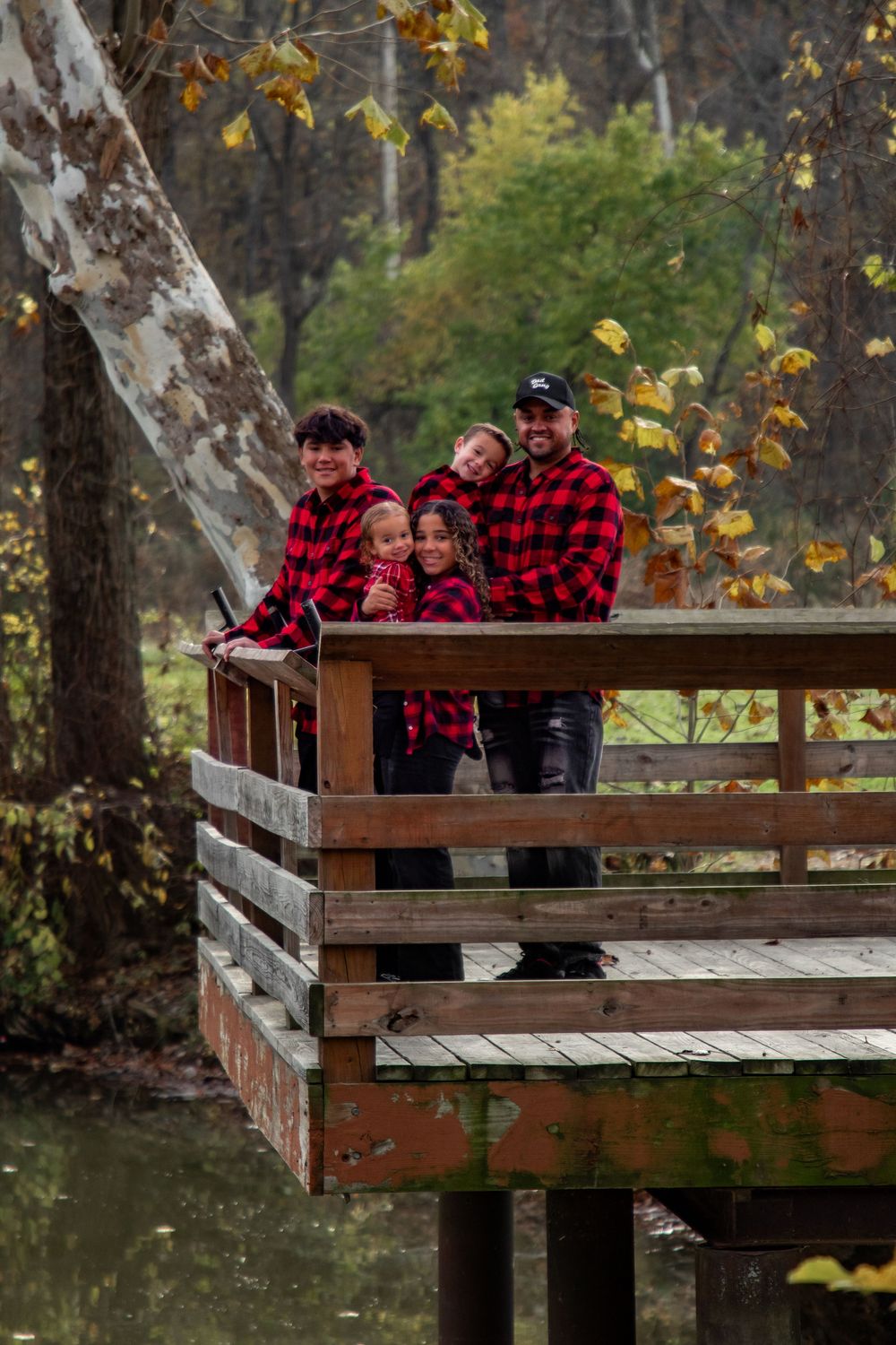 A family wearing matching red buffalo plaid shirts poses together on a wooden deck overlooking autumn trees.