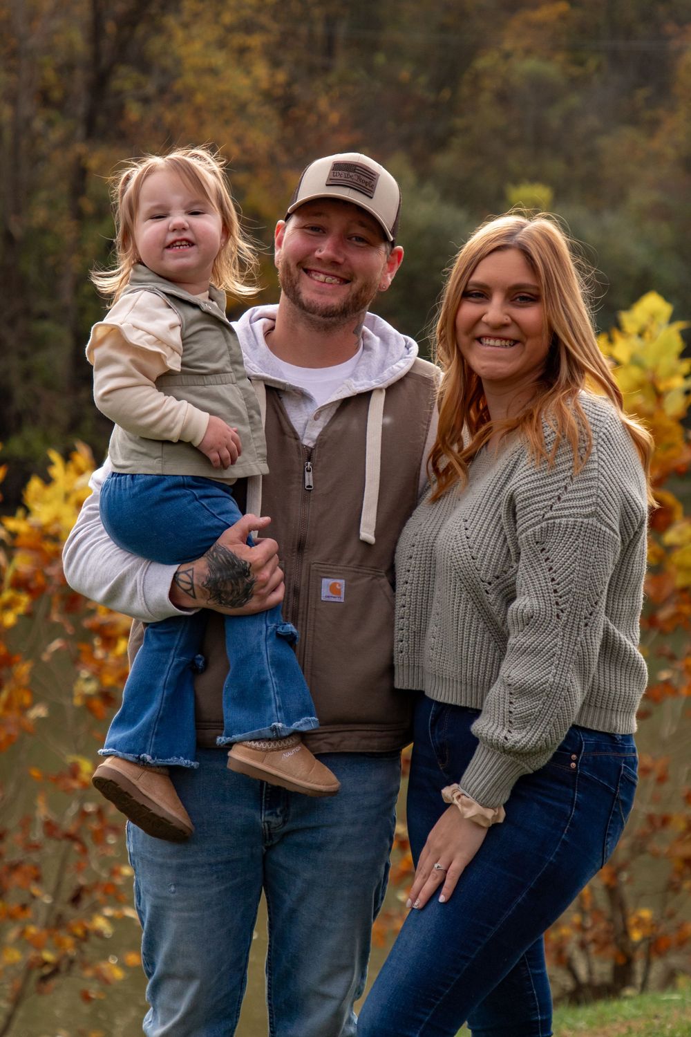 A family poses together outdoors in autumn wearing casual sweaters and jeans against a backdrop of fall foliage.