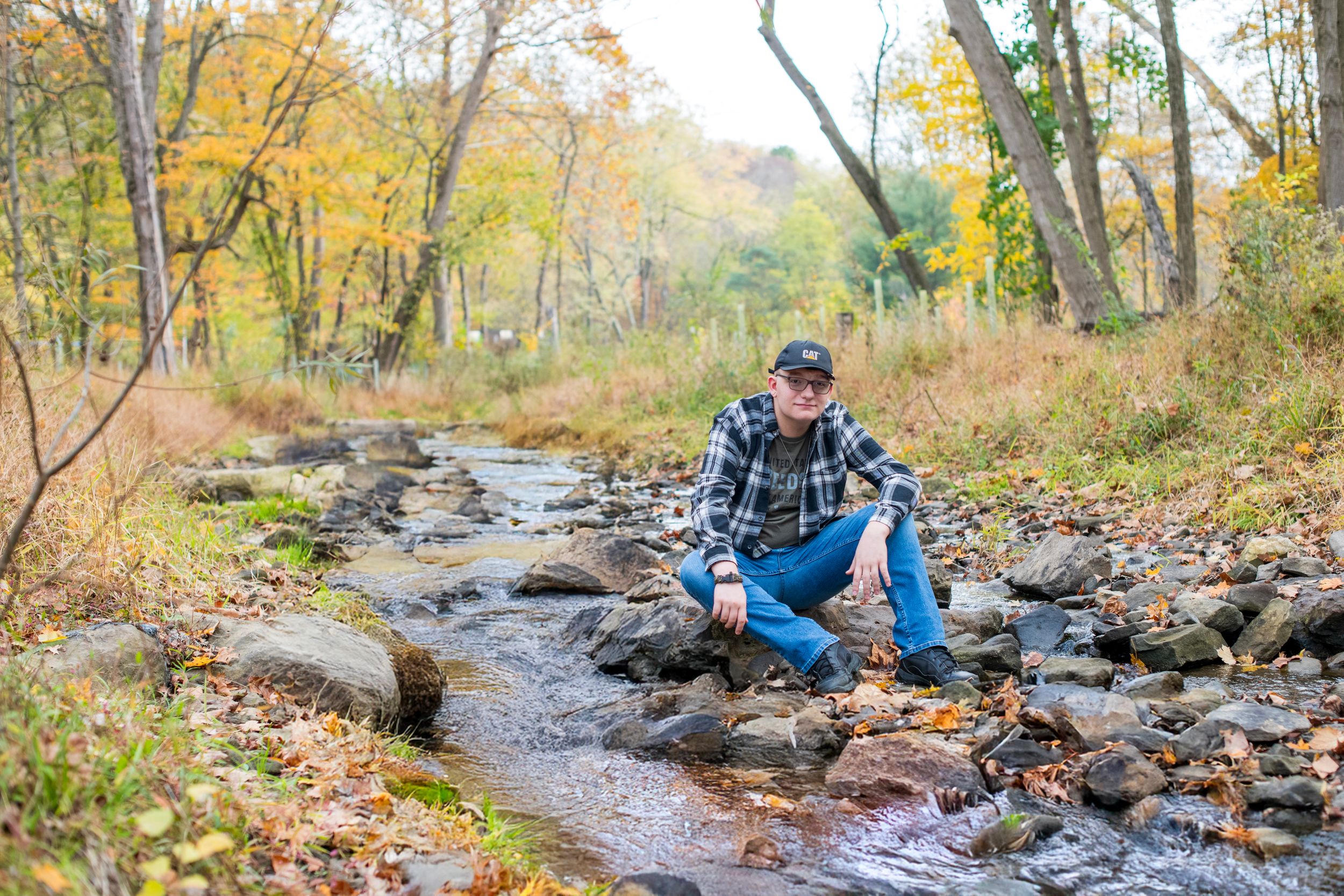 Man in plaid shirt and cap sitting on rocks beside a peaceful forest stream in autumn.