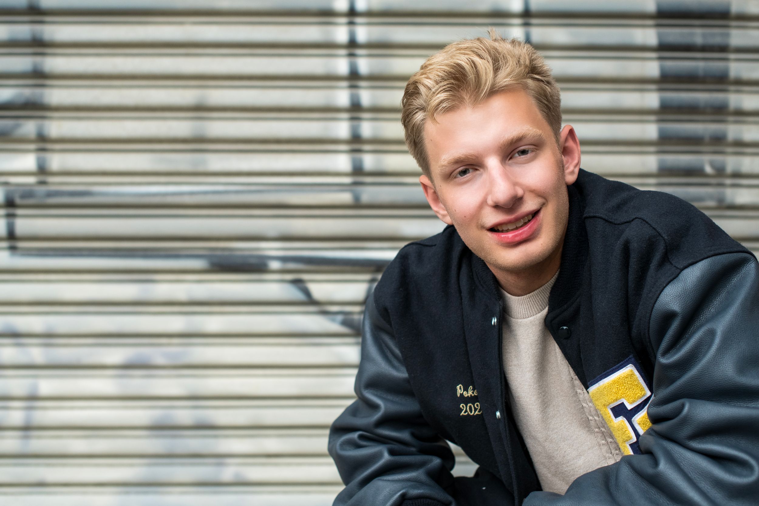Young blond man in varsity jacket smiling confidently in front of metal shutter backdrop.