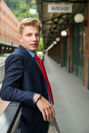 Young blond man in navy suit and red tie leaning on railing at McKnight train station platform.