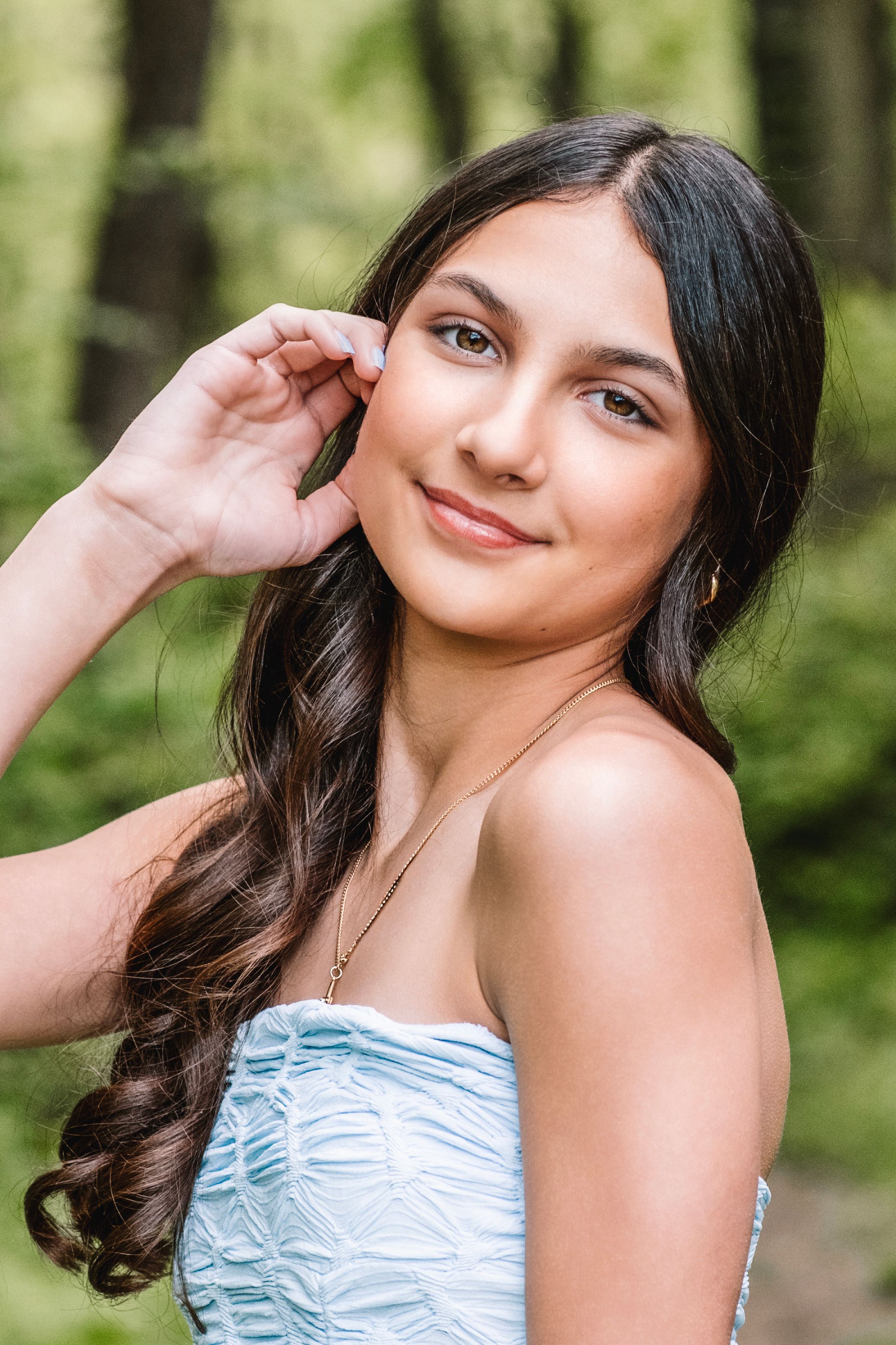 Young woman with dark wavy hair in a blue strapless dress smiling outdoors in a green forest setting.