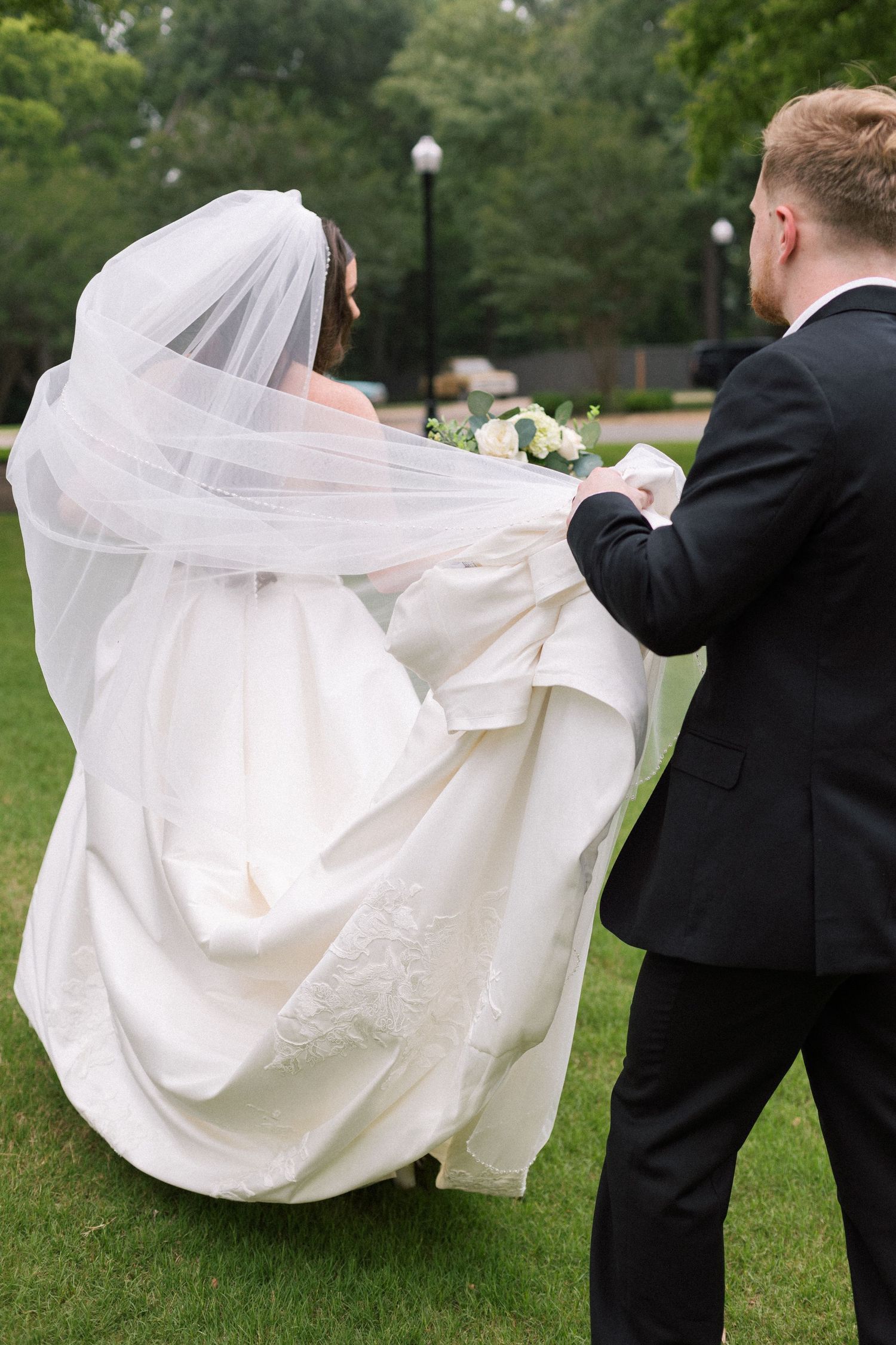 Groom in black suit helps bride in flowing white gown and veil walk across green garden lawn.