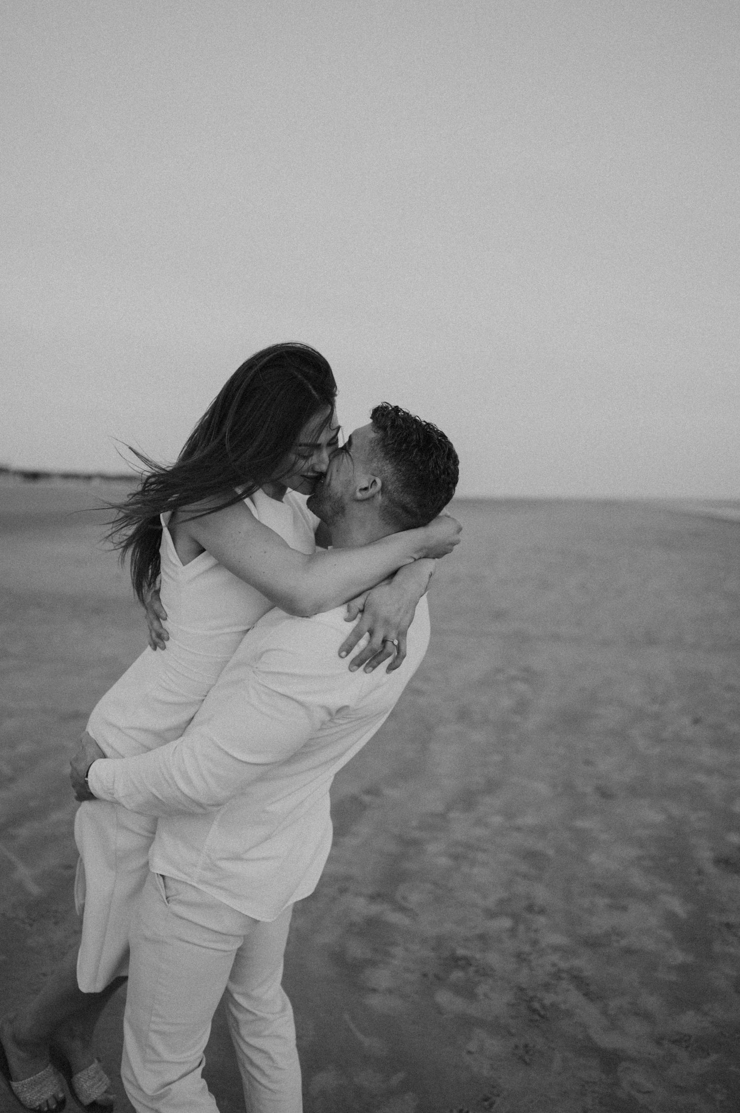 A romantic black and white photo series of a couple embracing on a beach in all-white attire.