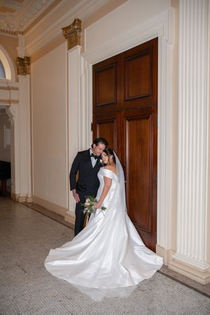 A wedding couple poses in front of a wooden door in a classic venue with white columns.