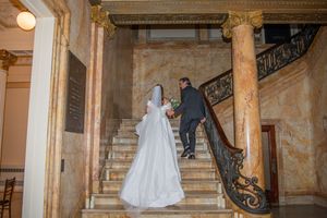 A wedding couple ascends an elegant marble staircase with ornate iron railings.