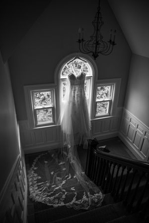 A dramatic black and white photo of a wedding dress hanging in a stairwell with natural light from arched windows.