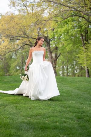 Bride in strapless white ball gown holding bouquet walks on lush green grass surrounded by spring trees.