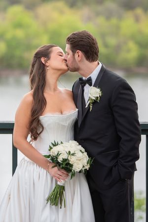 Bride and groom share a romantic kiss on a balcony, bride holding white rose bouquet in elegant strapless gown.