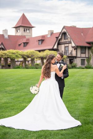 Bride in white gown and groom in tuxedo embrace on green lawn before Tudor-style mansion on wedding day.