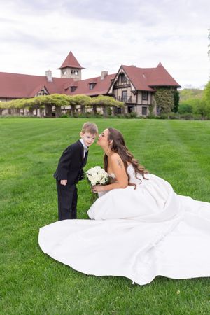 Bride in white gown kisses young boy in tuxedo on green lawn before elegant manor wedding venue.