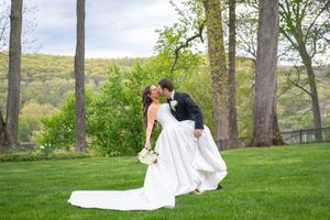 Bride and groom share a romantic dip kiss on lush green lawn with trees and rolling hills in background.